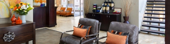 A modern hotel lobby with leather chairs, orange accent pillows, a coffee table with a plant, a cabinet with coffee dispensers, a TV on the wall, and large windows letting in natural light.