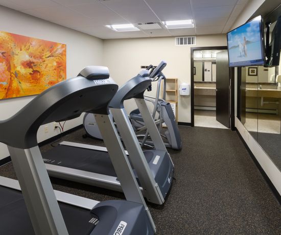 A small fitness room with two treadmills facing a wall-mounted TV. There is a large mirror on one wall, an abstract orange painting on another, and a paper towel dispenser near the door. The room has a speckled rubber floor.