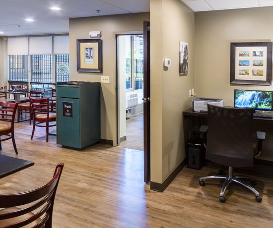 A small office area with a desk, chair, computer, printer, and lamp is next to a break room with tables and chairs. A doorway connects the spaces, and framed pictures decorate the beige walls.