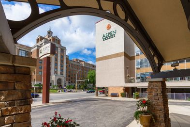 View from beneath a covered entryway showing the Aspen Suites hotel building and sign, with a larger multi-story building and partly cloudy blue skies in the background. Flowers and stone columns are in the foreground.