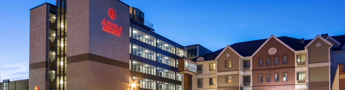 An exterior view of Aspen Suites Hotel at dusk, featuring illuminated signs and lights, a multi-story building on the left, and a smaller, gabled building on the right, with light trails from passing traffic in front.