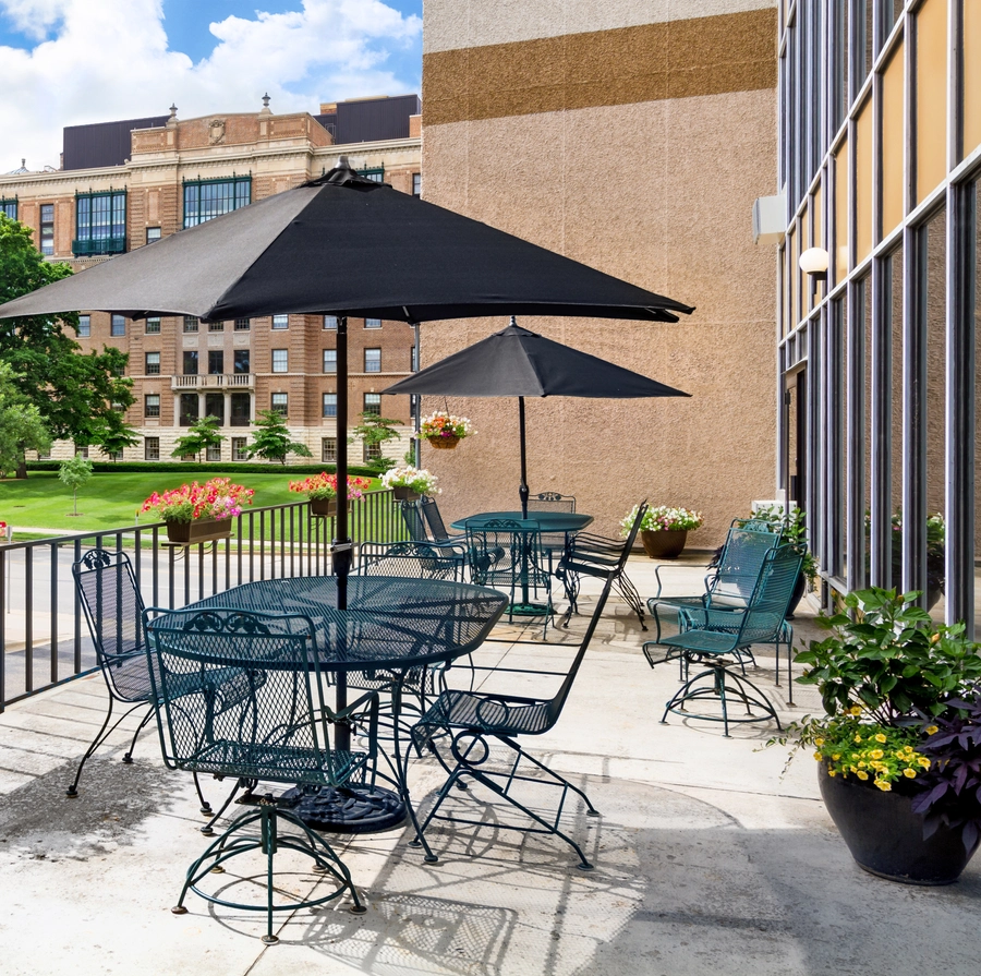 Outdoor patio with black metal tables and chairs, shaded by large black umbrellas. Potted flowers decorate the area, with a view of green lawn, trees, and brick buildings under a partly cloudy sky.