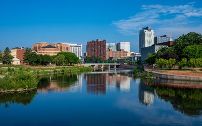 A city skyline with modern and historic buildings is reflected in a calm river, surrounded by green trees and blue sky with scattered clouds on a sunny day.